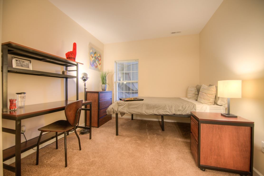 Bedroom in a University Edge apartment. There is a single bed against the exterior wall that has a small window. There is a nightstand with a lamp next to the bed. There is a desk with a shelf against the opposite wall.
